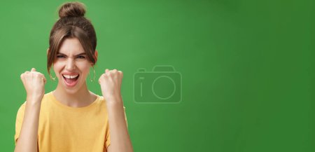Photo for Woman with spirit of winner raising clenched fists smiling excited and supportive cheering being ready for gym excercised boosing confidents with yell looking daring at camera over green wall - Royalty Free Image