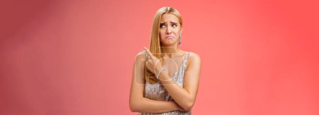 Photo for Upset doubtful unsure cute blond funny glamour girl in silver evening dress cringing grimacing hesitant pointing looking upper right corner suspicious, uncertain standing red background. - Royalty Free Image