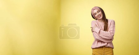Photo for Cozy charming friendly young smiling girl in knitted warm comfortable sweater, grinning joyfully tilt head emracing herself, crossing arms, hugging feeling safe and happy, posing against yellow wall. - Royalty Free Image