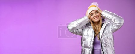 Photo for Tender romantic attractive blond female enjoying winter ski resort vacation having fun look pleased smiling broadly tilting head touching hat wearing silver stylish jacket, purple background. - Royalty Free Image