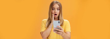 Photo for Waist-up shot of shocked and stunned emotive young woman reacting to shocking terrible news reading online from internet gasping opening mouth nervously touchin cheek staring at smartphone screen - Royalty Free Image