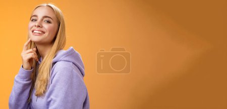 Téléchargez les photos : Attrayant sassy auto-assuré caucasien blond femelle en violet sweat à capuche tourner caméra toucher menton cheeky sourire caméra vérifier chose intéressante regardant vers l'avant intrigué, debout fond orane. - en image libre de droit