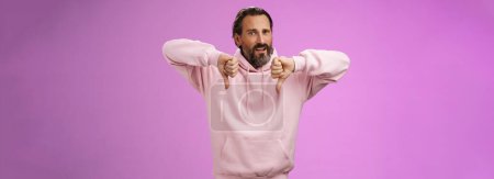 Photo for Disappointed upset angry caucasian adult bearded male show thumbs down dislking bad unprofessional behaviour frowning demanding refund standing bothered give negative feedback, purple background. - Royalty Free Image