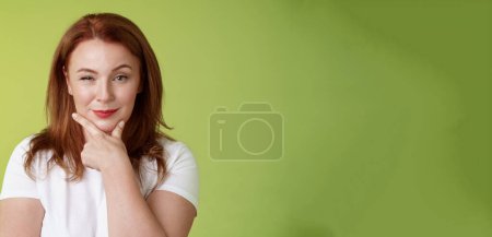 Téléchargez les photos : Choix intéressant. Intriguée rousse rusée femelle d'âge moyen tenir la main ligne du visage frotter menton souriant réfléchi heureux soulever sourcil curieux réfléchir décision séduisante fond vert. - en image libre de droit