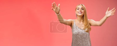 Photo for Friendly charming tender hospitable attractive birthday girl in luxury silver dress welcoming guests throw party extend arms cuddle greeting people hug give cuddle smiling broadly, red background. - Royalty Free Image