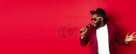 Photo for Passionate black male singer performing against red background, singing into microphone, wearing party outfit, standing over red background. - Royalty Free Image