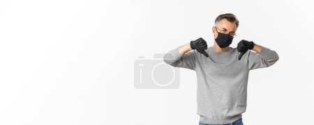 Photo for Concept of coronavirus, lifestyle and quarantine. Portrait of unamused and skeptical middle-aged man dislike something, wearing medical mask and gloves, showing thumbs-down. - Royalty Free Image