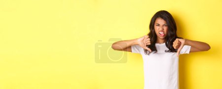 Photo for Portrait of disappointed afrcan-american teenage girl, showing tongue from dislike and thumbs-down, judging something bad, standing over yellow background. - Royalty Free Image
