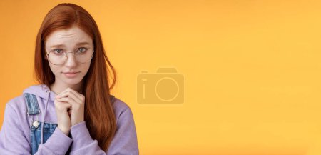 Photo for Silly guilty young shy redhead girlfriend asking forgiveness supplicating lower head look from under forehead frowning begging apology favour standing insecure sad pleading help, orange background. - Royalty Free Image