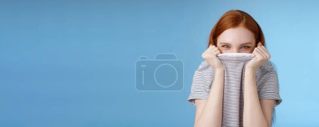Photo for Silly flirty amused attractive playful redhead girlfriend hiding face pulling t-shirt head squinting devious mysteriously giggle laughing hope disguise pranking friend standing blue background. - Royalty Free Image