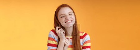 Photo for Happy dreamy romantic young tender ginger girl fantasizing creating love story imagination smiling broadly delighted close eyes touching hair strands recalling nice memory, standing orange background. - Royalty Free Image