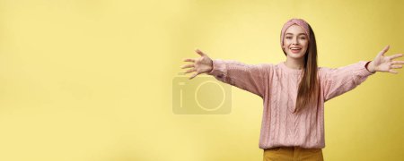 Foto de Atractiva amigable joven europea positiva con suéter, diadema de bienvenida brazos extanding y sonriendo a la cámara dando abrazos, abraza amigos, abrazos contra el fondo amarillo. Copiar espacio - Imagen libre de derechos