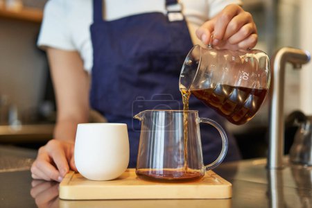 Téléchargez les photos : Gros plan du bocal en verre et d'une tasse debout sur le comptoir du café, barista versant du café filtre et préparant l'ordre. - en image libre de droit