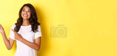 Photo for Portrait of beautiful african-american woman, smiling and looking dreamy at promo, pointing fingers at upper left corner, showing logo over yellow background. - Royalty Free Image