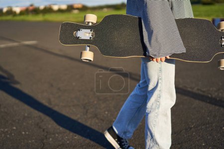 Téléchargez les photos : Coupé coup de jeune fille patineuse, mains tenant longboard, marche avec planche à roulettes sur route en béton. - en image libre de droit