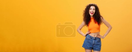 Stylish confident and awesome woman. ready lit party standing in trendy cropped top and denim shorts with hands on waist sticking out tongue and smiling joyfully posing over orange background.