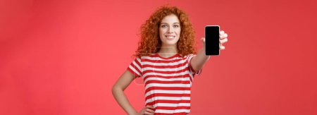Photo for Confident good-looking redhead curly woman present cool app hold smartphone extended hand show phone screen smiling assertive recommending subscribe her blog social-media. Copy space - Royalty Free Image