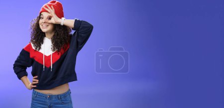 Photo for Okay girl agrees. Friendly-looking happy and charming young curly-haired girl in warm beanie showing ok gesture or circle over eye as peeking at camera and smiling broadly with joy over blue wall. - Royalty Free Image