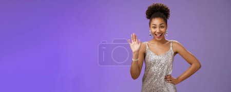 Photo for Friendly elegant african-american young 20s woman. wearing elegant silver glittering dress hold hand waist waving hello greeting gesture say hi informally meet friend talking blue background. - Royalty Free Image