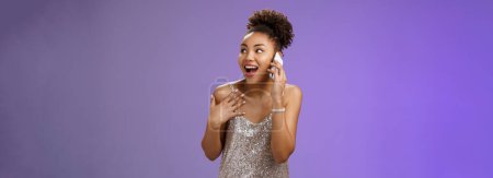 Photo for Impressed sociable african-american talkative woman in silver dress talking smartphone pointing herself amused describing emotions impressions after visit awesome party, blue background. - Royalty Free Image
