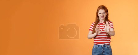 Photo for Gosh it stinks. Disgusted redhead picky woman blocking sign raise hands up defensive, grimacing, cringe from aversion awful smell, show refusal rejecting disgusting offer, stand orange background. - Royalty Free Image