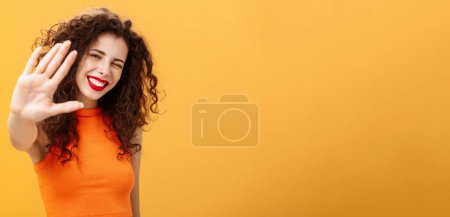 Photo for Give me five mate. Stylish friendly and joyful attractive woman with curly hairstyle winking and smiling happily pulling hand towards camera to greet or congratulate friend over orange background - Royalty Free Image