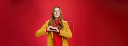 Photo for Love you all. Portrait of romantic and stylish good-looking flirty redhead female with freckles and blue eyes folding lips to give kiss showing heart gesture, confessing in sympathy over red wall. - Royalty Free Image