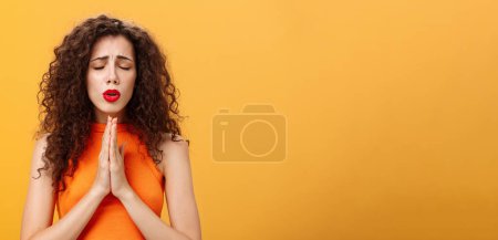 Photo for Nervous and concerned woman. with curly hairstyle feeling hopeful praying with closed eyes and frowned eyebrows holding hands in pray near chest hopefully dreaming troubled will solve over orange wall - Royalty Free Image