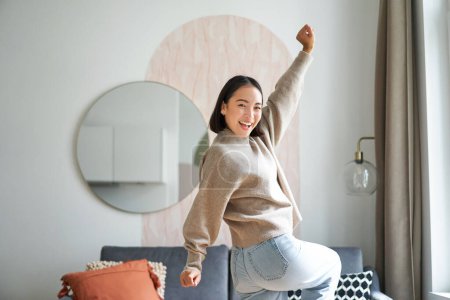 Foto de Linda mujer asiática emocionada bailando, levantando las manos en triunfo, celebrando y cantando, de pie en casa en tibio jamper. - Imagen libre de derechos