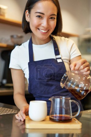 Téléchargez les photos : Barista dans le café versant le café filtre, brassant et préparant l'ordre derrière le comptoir, portant le tablier bleu. - en image libre de droit