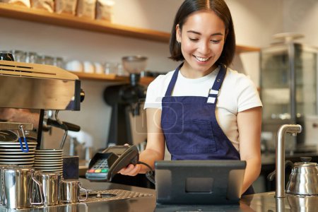 Lächelnde asiatische Barista, die Bestellung bearbeitet, Bestellung im Kassenterminal eingibt, am Schalter im Café arbeitet.