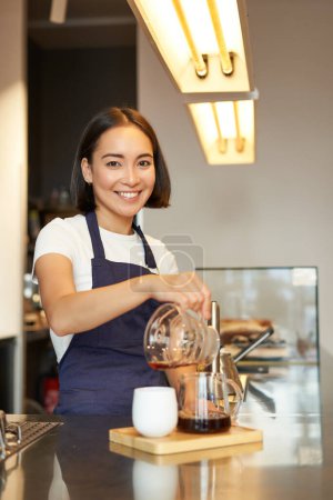 Téléchargez les photos : Plan vertical de fille souriante travaillant comme barista, préparer verser plus, fabrication de café filtre mélange, debout au comptoir dans le café dans le tablier. - en image libre de droit