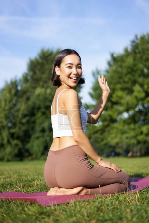 Téléchargez les photos : Portrait de fille asiatique en cours de fitness dans le parc, assis sur tapis de caoutchouc et main vague à la caméra, dire bonjour, méditation et yoga pratique. - en image libre de droit