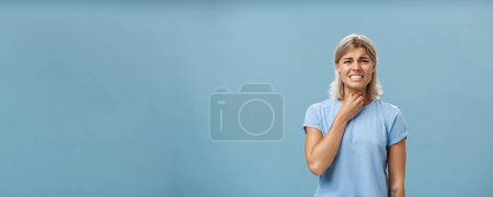 Photo for Girl feeling discomfort in throat catching cold or having seasonal allergy touching neck and frowning with clenched teeth and painful expression standing displeased over blue background. Health - Royalty Free Image