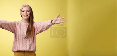 Foto de Atractiva amigable joven europea positiva con suéter, diadema de bienvenida brazos extanding y sonriendo a la cámara dando abrazos, abraza amigos, abrazos contra el fondo amarillo. Copiar espacio - Imagen libre de derechos