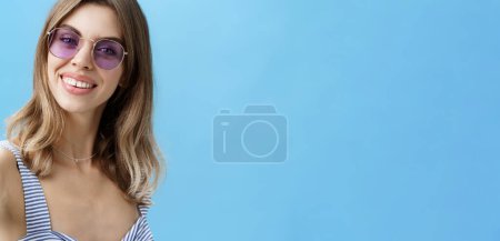 Téléchargez les photos : Charmante femme avec de jolies dents bénies dans des lunettes de soleil se détendre en vacances en prenant de beaux clichés tirant la main vers la caméra le tenant pour prendre selfie souriant largement sur fond bleu. Mode de vie. - en image libre de droit