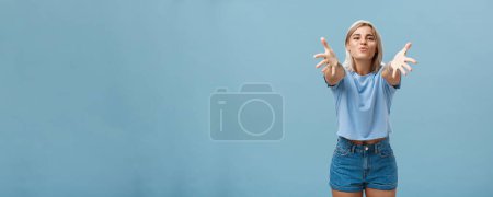 Photo for Come to mama. Portrait of friendly joyful and enthusiastic caucasian female student in trendy outfit pulling hands towads camera and folding lips to hug and give passionate kiss smiling over blue wall - Royalty Free Image