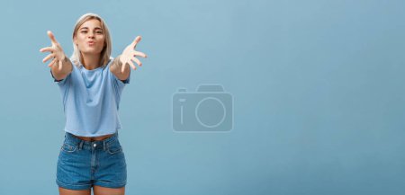 Photo for Come to mama. Portrait of friendly joyful and enthusiastic caucasian female student in trendy outfit pulling hands towads camera and folding lips to hug and give passionate kiss smiling over blue wall - Royalty Free Image