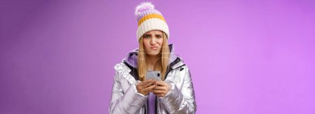 Photo for Perplexed displeased cute blond stylish girl unwilling anser message cringing reluctant frowning holding smartphone receive disappointed perplexing text, standing unsure purple background. - Royalty Free Image