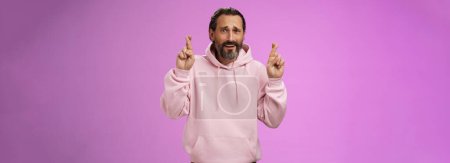 Photo for Nervous unsure hopeful handsome bearded 40s man in pink stylish hoodie cringing worried cross figers good luck make wish hope not gonna lose job supplicating praying dream come true. Lifestyle. - Royalty Free Image