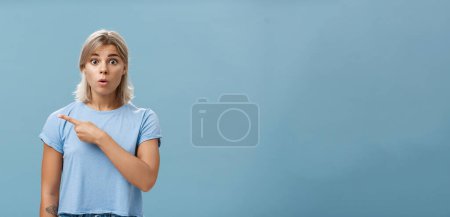 Photo for Studio shot of amazed stunned cute blonde witnessing unbelievable event gasping opening mouth staring astonished and pointing left being questioned and shocked over blue background. Emotions concept - Royalty Free Image