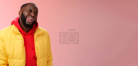 Téléchargez les photos : Cheeky élégant drôle homme barbu africain en veste jaune à la mode sweat à capuche rouge montrer la langue sassy flirt regard clin d'oeil caméra essayer d'impressionner femme feindre macho, debout fond rose. - en image libre de droit