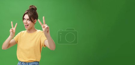 Photo for Indoor shot of enthusiastic excited and happy daring girl with combed hair tattoo and cute diasdema showing peace signs bending backwards standing in cool energized pose over green background - Royalty Free Image
