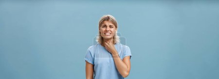 Photo for Girl feeling discomfort in throat catching cold or having seasonal allergy touching neck and frowning with clenched teeth and painful expression standing displeased over blue background. Health - Royalty Free Image