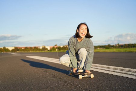 Téléchargez les photos : Liberté et bonheur. Heureuse fille asiatique chevauchant son longboard sur une route ensoleillée vide, riant et souriant, skateboard. - en image libre de droit