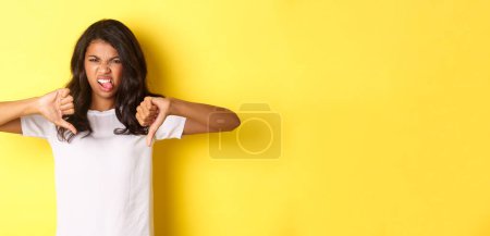 Photo for Portrait of disappointed afrcan-american teenage girl, showing tongue from dislike and thumbs-down, judging something bad, standing over yellow background. - Royalty Free Image