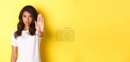 Foto de Imagen de mujer afroamericana confiada y seria, diciendo no, mostrando señal de stop y frunciendo el ceño disgustado, prohibiendo algo malo, de pie sobre fondo amarillo. - Imagen libre de derechos