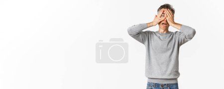 Téléchargez les photos : Image de embarrassé de l'homme d'âge moyen effrayé couvrir ses yeux avec les mains, regardant à travers les doigts avec une expression choquée, debout sur fond blanc. - en image libre de droit