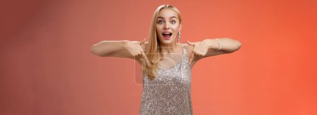 Photo for Impressed excited attractive glamour blond girl in silver glittering dress gasping thrilled pointing down glance camera fascinated check out fabulous awesome promo, standing amused red background. - Royalty Free Image