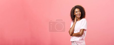 Foto de Amor en el aire ven y saluda. Coqueta sensual y atractiva joven africana americana estudiante con peinado afro tocando el cuello girando suavemente a la derecha, mirando con curiosidad y deseo sobre la pared rosa. - Imagen libre de derechos
